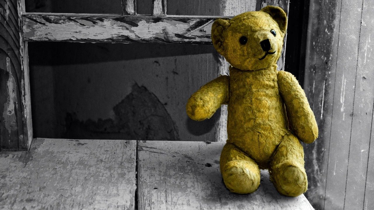 A small worn golden teddy bear with jointed arms sitting on a weathered wooden ledge in front of a gray window frame