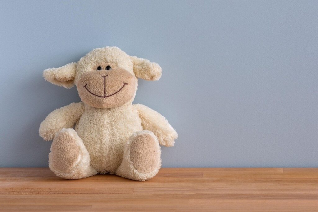 Cream-colored plush lamb with a stitched smile sitting on a wooden shelf against a pale blue wall