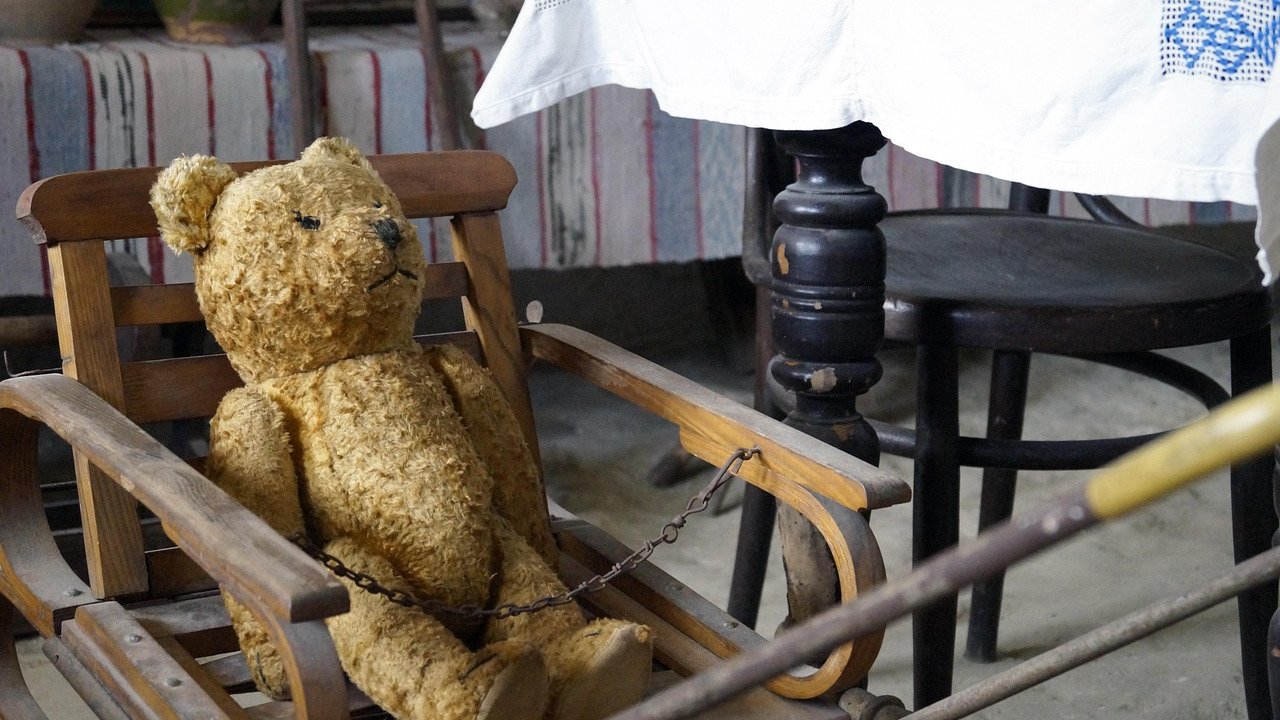 A worn golden mohair-style teddy bear sitting in a small old wooden chair with a chain across its lap
