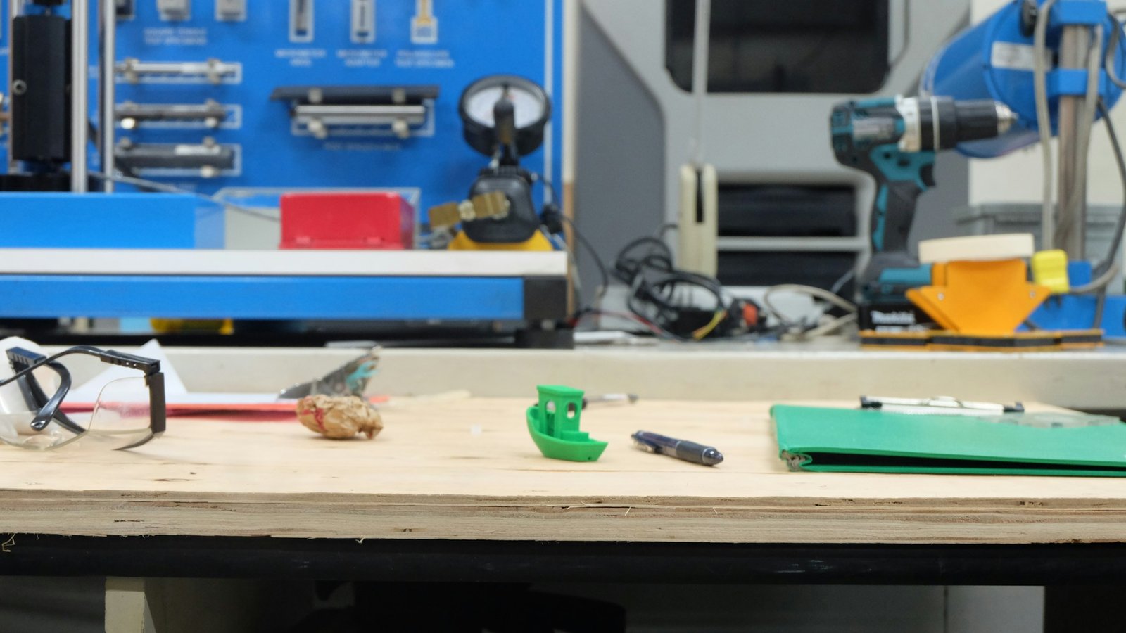 Workshop bench with a small green plastic prototype part, a pen, safety glasses and power tools in the background