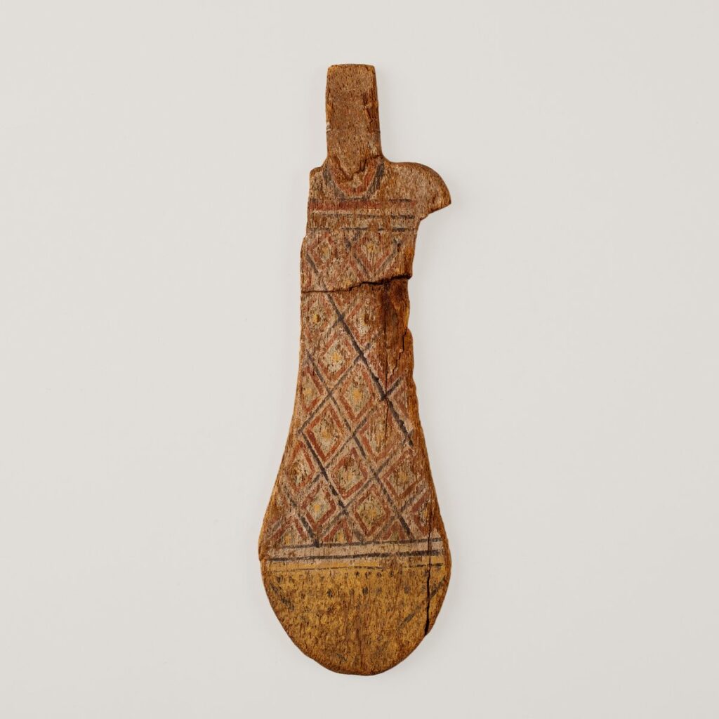 Keyhole-shaped wooden paddle doll painted with a diamond-patterned garment and a necklace, missing its bead hair