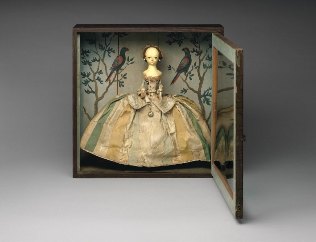 Wooden English fashion doll in a wide silk skirt standing inside a painted shadow box decorated with trees and parrots