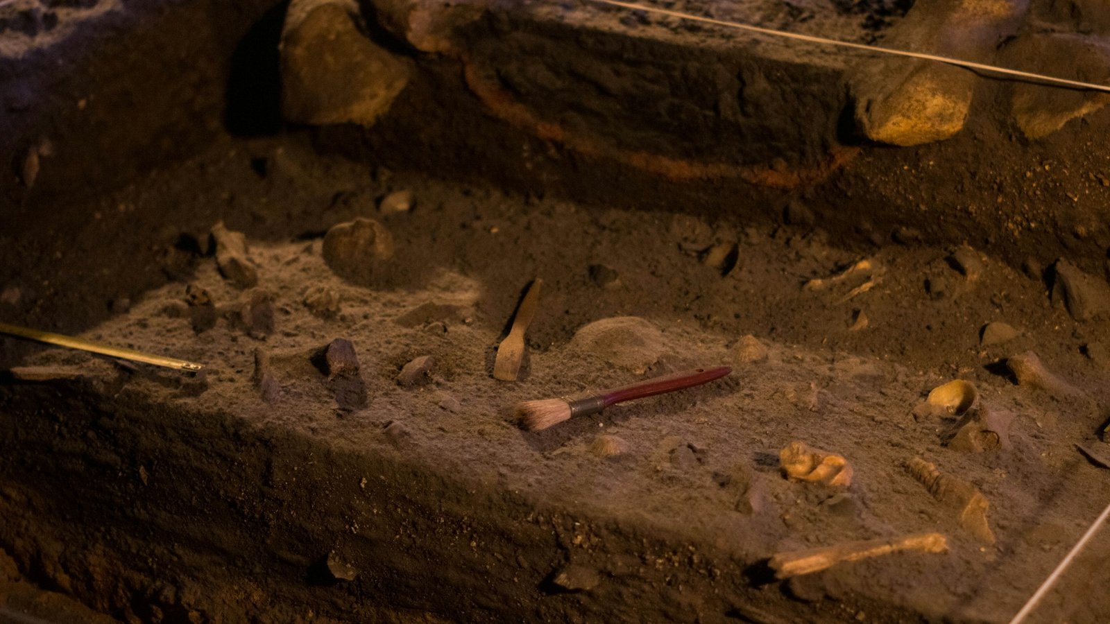 Shallow excavation trench with exposed stones and a red-handled brush resting in the soil