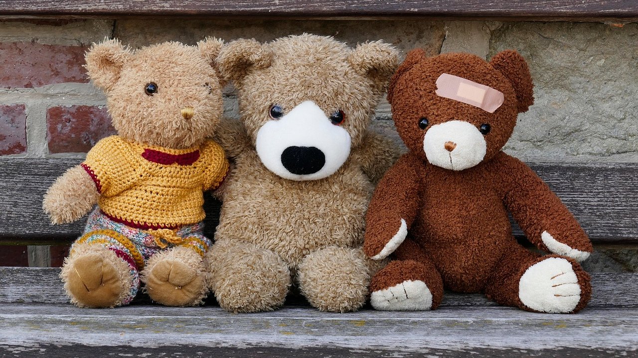 Three well-loved teddy bears, one wearing a knitted sweater and one with a bandage, sitting on a weathered wooden bench