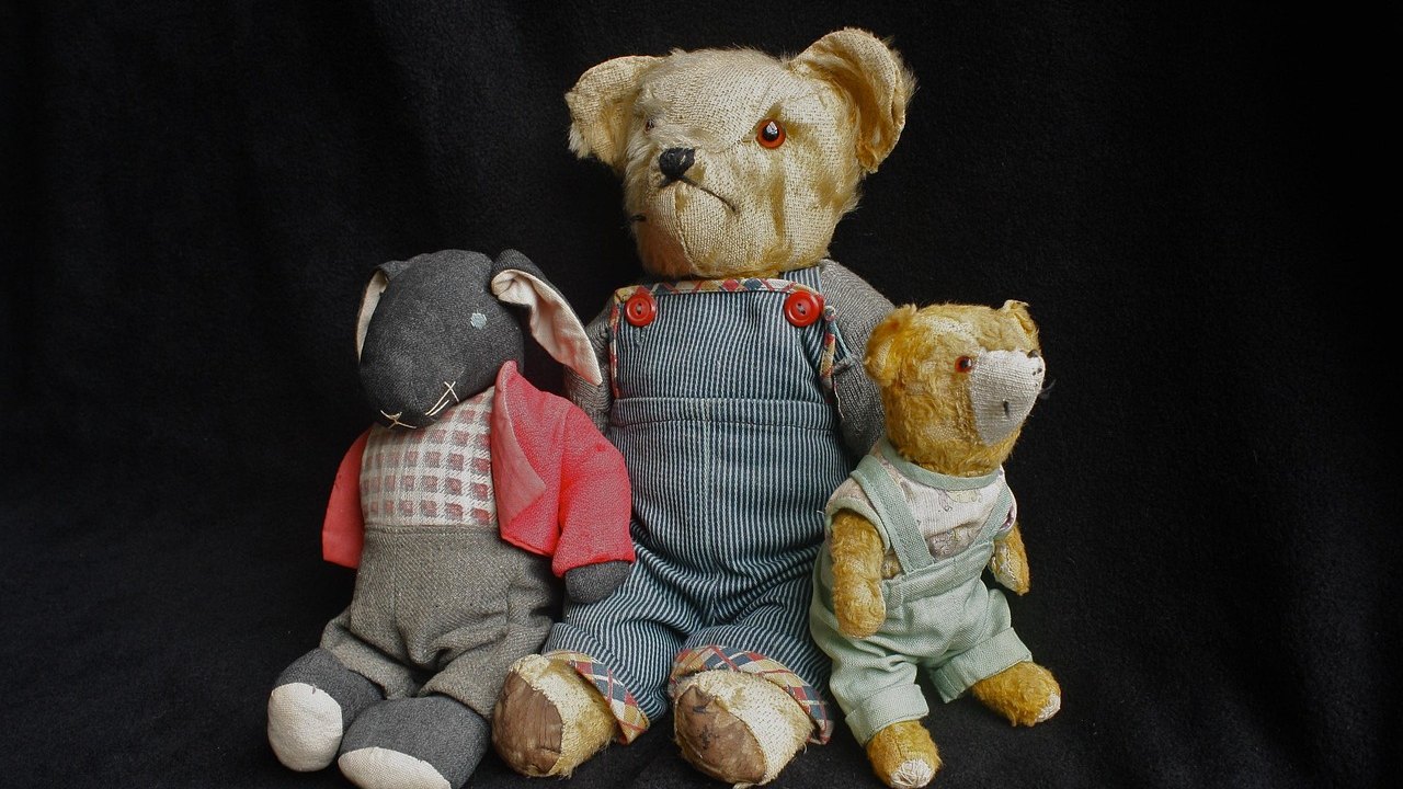 Three old stuffed toys, a worn bear in overalls, a smaller mohair bear, and a cloth mouse, against a dark background