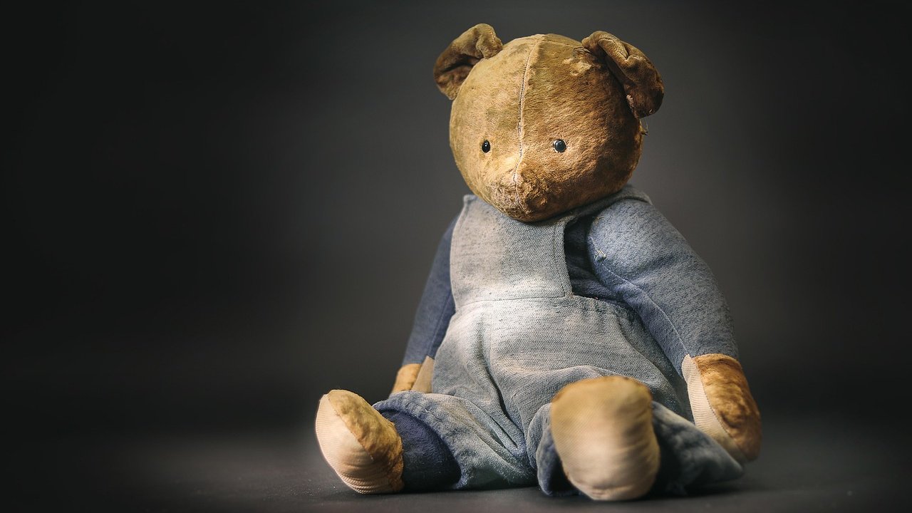 A worn old teddy bear in faded blue overalls sitting against a dark background