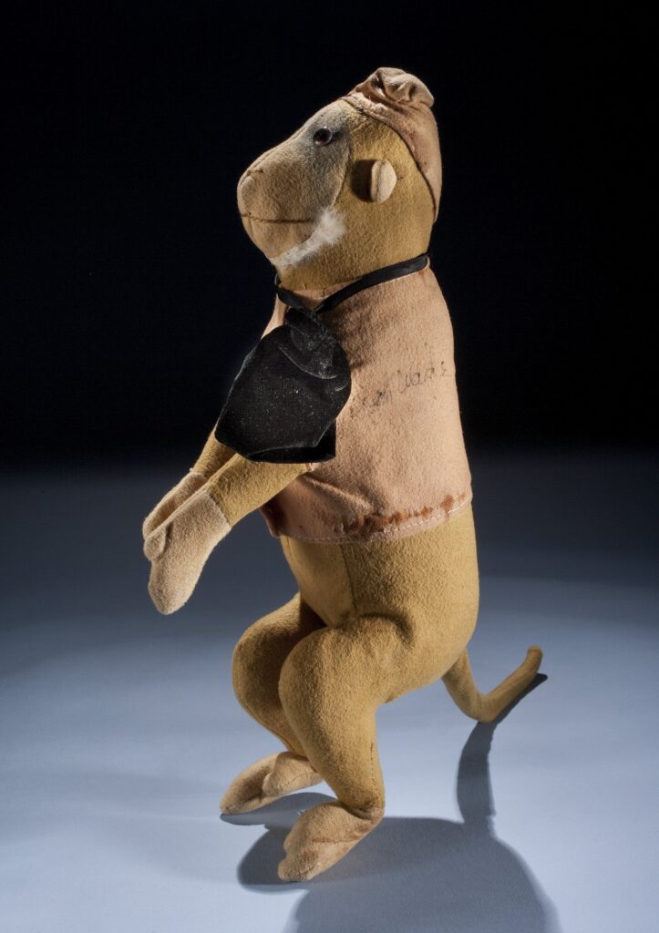 Tan cotton stuffed toy monkey standing upright, wearing a light vest, a small cap, and a black drawstring at the neck