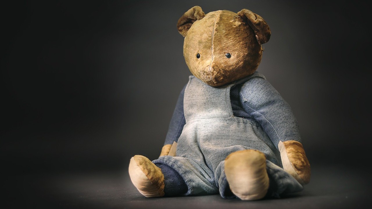 A worn, old mohair-style teddy bear with faded fur, dressed in blue overalls, sitting against a dark background