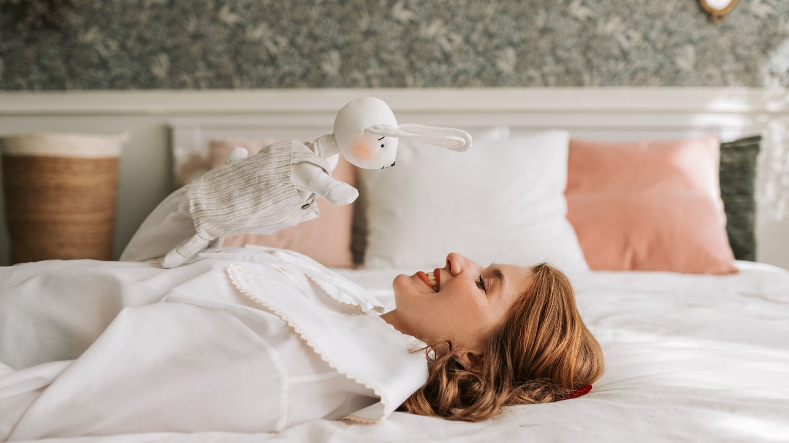 A smiling woman lying on a white bed holding up a soft white plush bunny