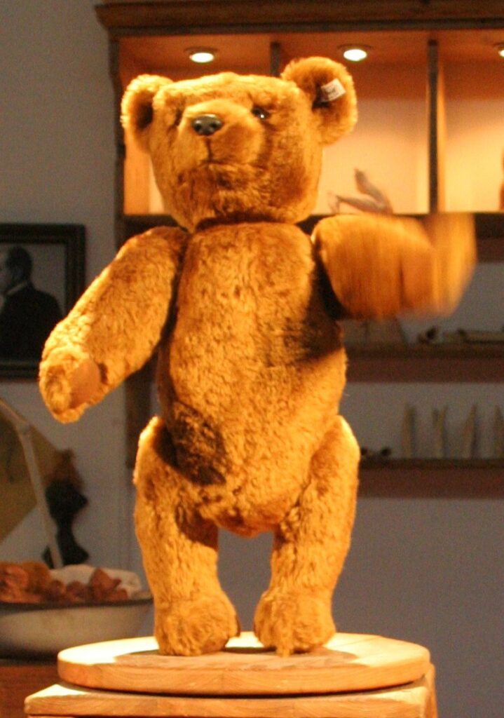 Modern replica of the Steiff Bear 55 PB, a tall jointed mohair bear with long arms, displayed on a wooden stand in the Steiff Museum in Giengen