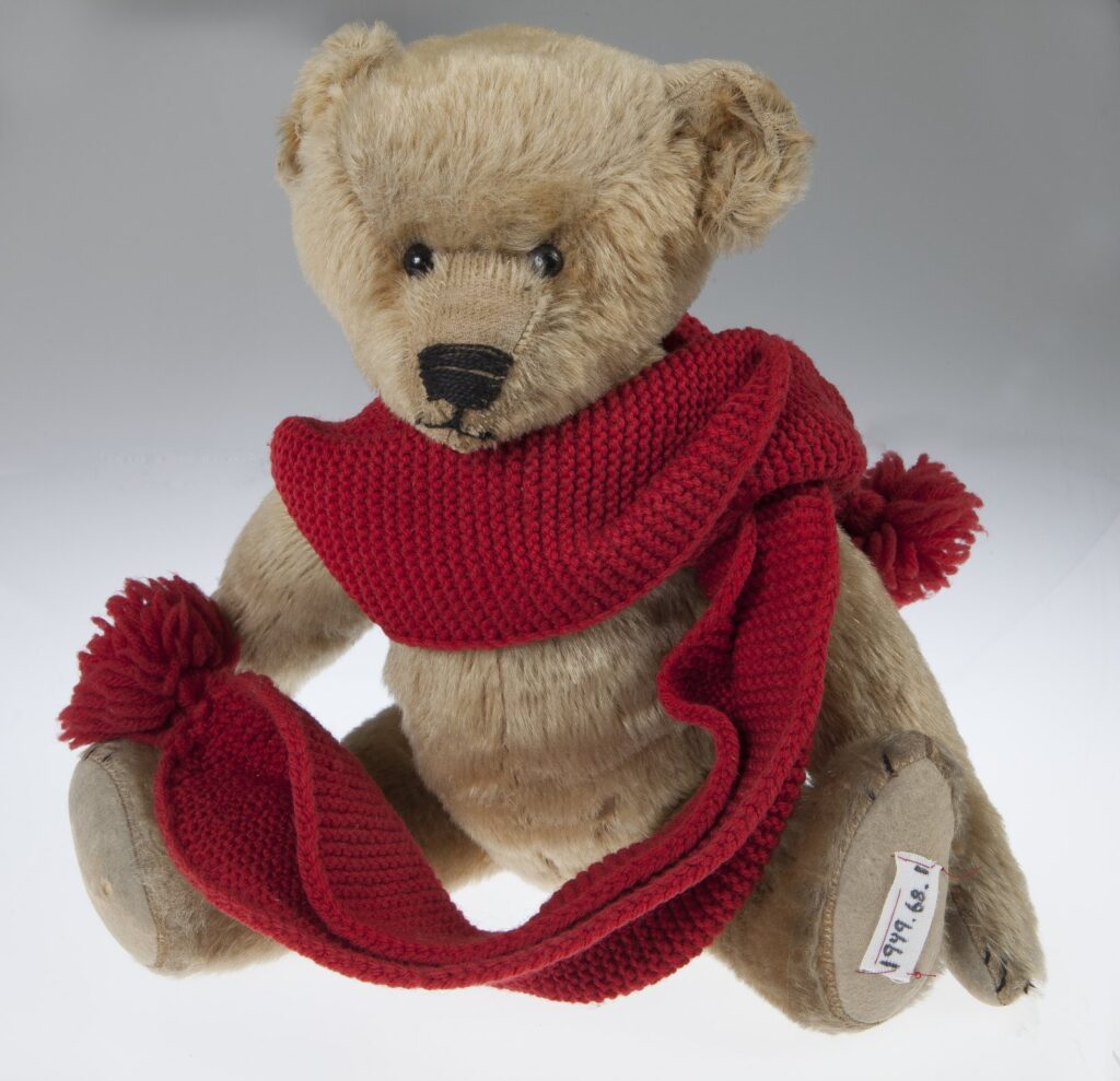 Beige mohair teddy bear with shoe-button eyes, a stitched nose, and a red knitted scarf, photographed against a white background