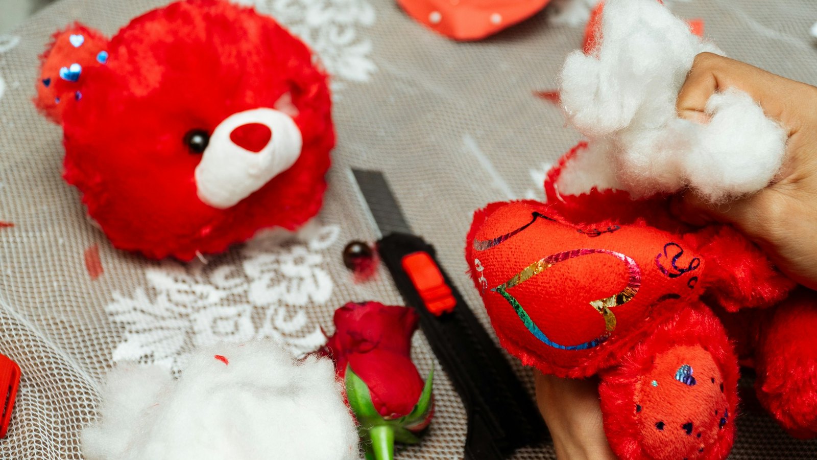 Hands pushing white fiber filling into a red plush bear and heart-shaped plush pieces on a work table, with a craft knife nearby