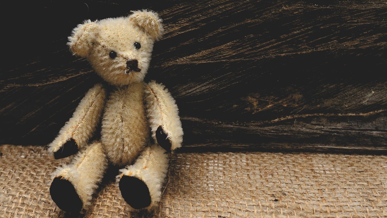 A worn vintage jointed teddy bear sitting on burlap against a dark wooden background