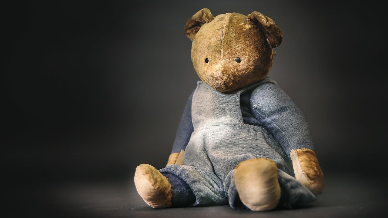 A well-worn old teddy bear in faded blue overalls sitting against a dark gray background