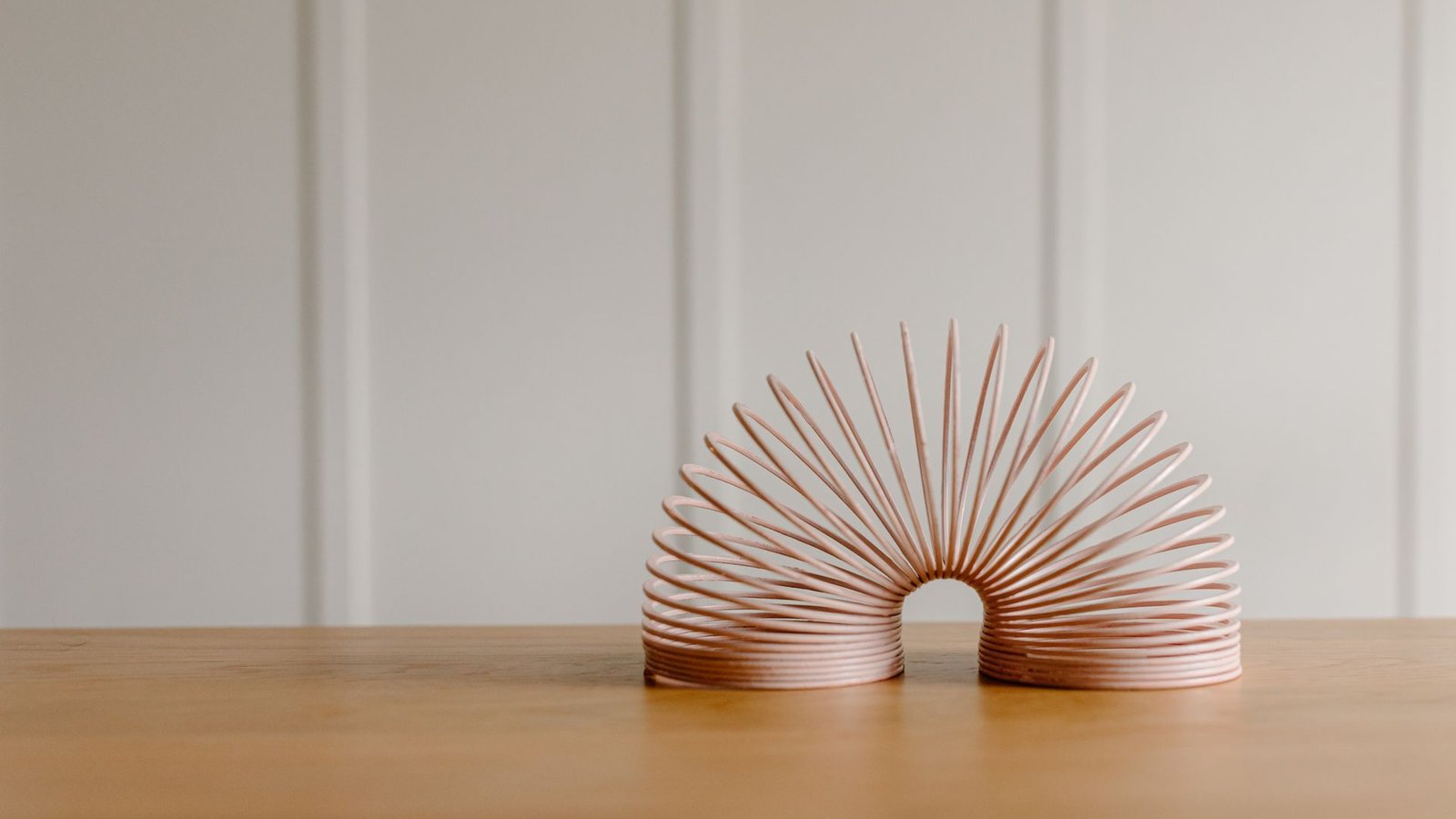 Pale pink coiled metal spring toy arched on a wooden table against a white paneled wall
