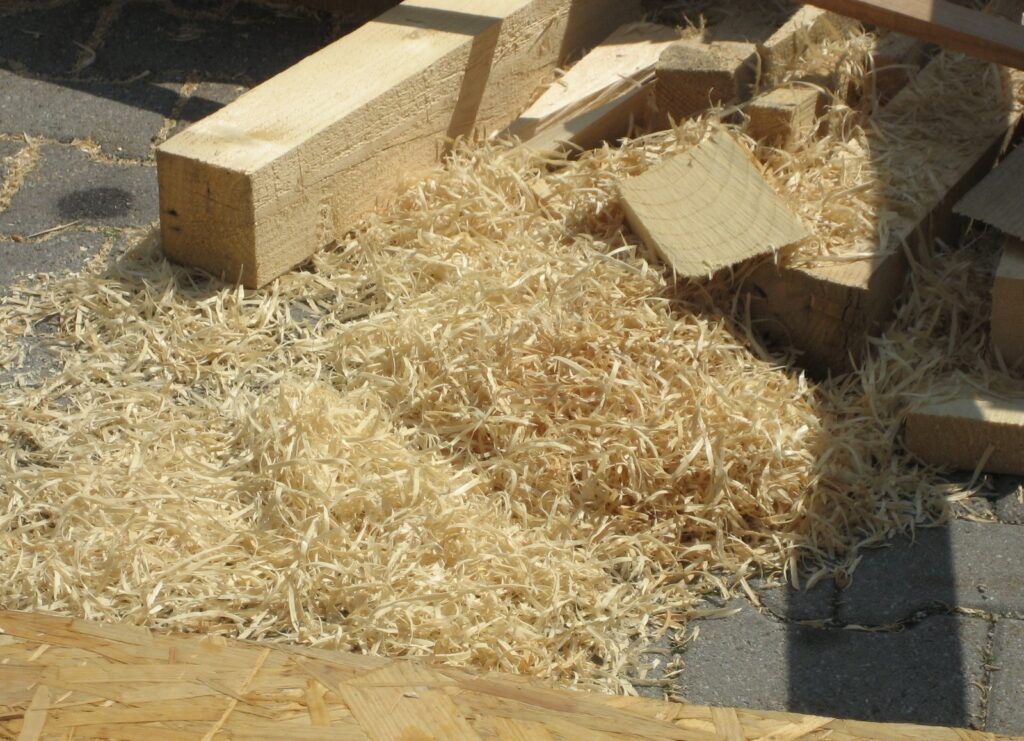 A pile of loose, pale wood wool shavings on the ground beside cut pieces of timber